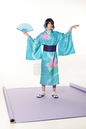 Ausdrucksstarker Mann in blauem Kimono und Perücke posiert mit Handfächer auf lila Teppich und weißem Hintergrund