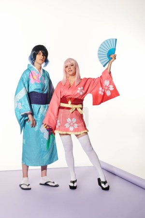 mujer de estilo anime posando con ventilador de mano cerca de hombre en kimono y peluca en alfombra púrpura en estudio blanco