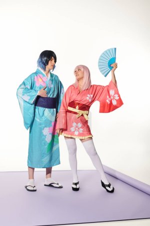 mujer rubia con peluca de mano cerca del hombre en kimono colorido en la alfombra púrpura en blanco, cultura cosplay