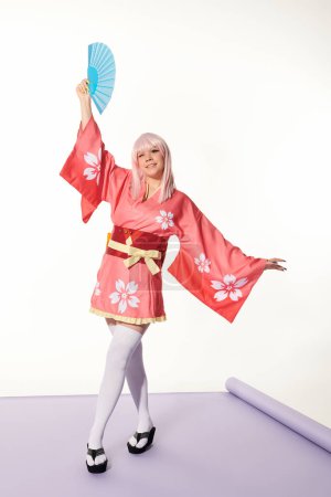 encantadora mujer de estilo anime en kimono rosa y peluca de pie con ventilador de mano en pose artística sobre blanco