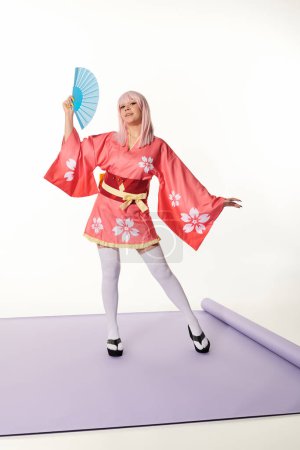 künstlerische Cosplay-Frau in rosa Kimono und blonder Perücke mit Handfächer auf lila Teppich im weißen Studio