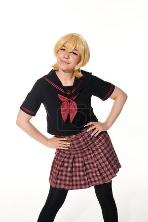 jeune femme ludique en perruque blonde jaune et uniforme scolaire avec les mains sur les hanches sur blanc, cosplay