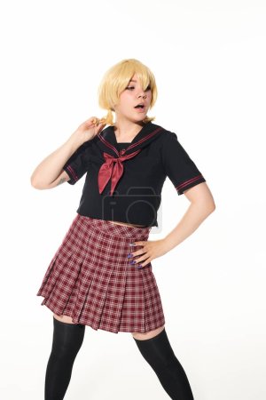 überraschte Anime-Frau in Schuluniform und gelbblonder Perücke, die mit der Hand an der Hüfte auf weiß steht