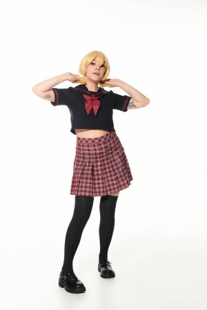 tattooed playful anime woman in school uniform touching ponytails on yellow blonde wig on white