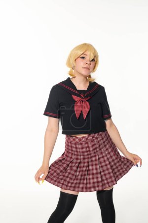 young anime style woman in yellow blonde wig posing in plaid skirt ant looking at camera on white