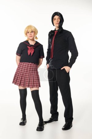 man in black clothes with hands in pockets near blonde woman in school uniform on white, cosplayers