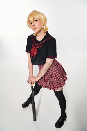 longitud completa de la mujer del anime en uniforme escolar de moda con bate de béisbol mirando a la cámara en blanco