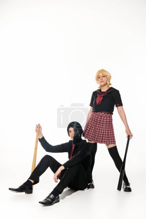 Téléchargez les photos : Couple confiant de style anime en uniforme scolaire et perruques avec posant avec des chauves-souris de baseball sur blanc - en image libre de droit