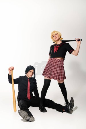 man in black wig sitting  with baseball bat near woman in school uniform on white, cosplay trend