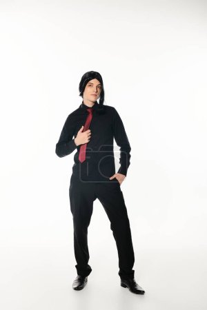 junger Cosplayer in schwarzer Kleidung und roter Krawatte, die Hand in der Tasche auf weiß, Jugendkultur