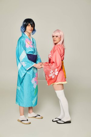 pareja joven en coloridos kimonos tomados de la mano y mirando a la cámara en gris, concepto de anime