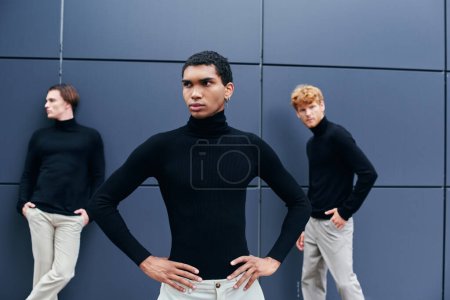 hombre afroamericano guapo en cuello alto negro con pendiente posando delante de otros hombres