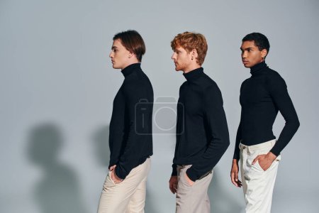 tres hombres multiétnicos en cuellos altos negros caminando en una sola fila sobre fondo gris, concepto de moda