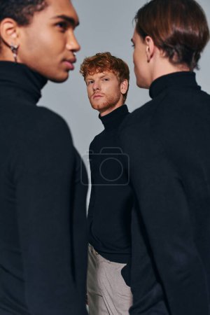 enfoque en el hombre joven de pie cerca borrosa amigos interracial sobre fondo gris, moda, vertical