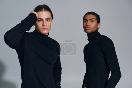 elegante interrassische Männer posieren an grauer Wand in schwarzen Rollkragenpullovern, Hand in Gesicht, Accessoires