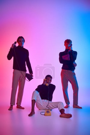 vertical shot of diverse trio in black attire surrounded by neon lights, landline phones, men power