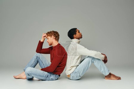 Téléchargez les photos : Deux jeunes hommes multiethniques dans des pulls chauds confortables assis sur le sol dos à dos, concept de mode - en image libre de droit