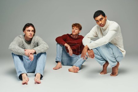 joven afroamericano hombre en suéter en cuclillas con otros dos hombres sentados en el suelo, concepto de moda