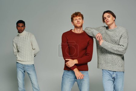 two young men in sweaters posing with their african american friend on backdrop, fashion concept