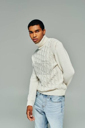 atractivo hombre afroamericano en suéter blanco de pie y mirando a la cámara, la mano detrás de la espalda