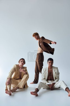 vertical shot of three multicultural men in elegant suits posing lively on gray background, fashion