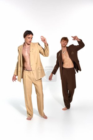 vertical shot of good looking elegant duo in unbuttoned suits posing barefoot in motions, men power