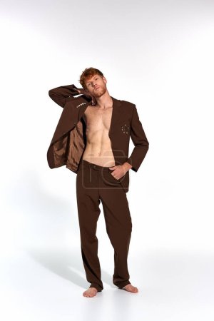 appealing red haired man in unbuttoned classy suit with accessories with hand behind head, fashion