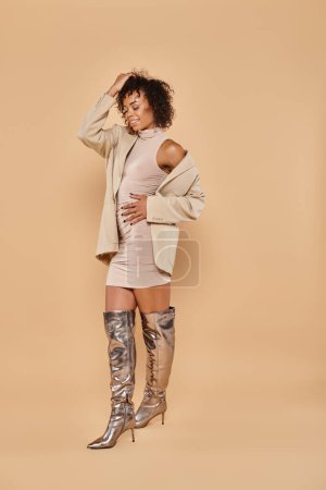 full length of african american woman with curly hair posing in autumnal attire on beige background