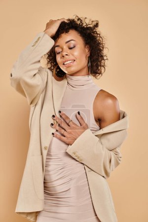 joyful african american woman with curly brunette hair posing in autumnal attire on beige background