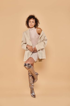 full length of stylish african american woman with curly hair posing in autumn outfit on beige