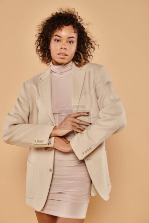 Photo for Beautiful african american woman with curly hair posing in autumn dress and blazer on beige backdrop - Royalty Free Image