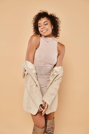Herbst-Trends, fröhliche Afroamerikanerin posiert in stylischem Blazer und Kleid auf beigem Hintergrund
