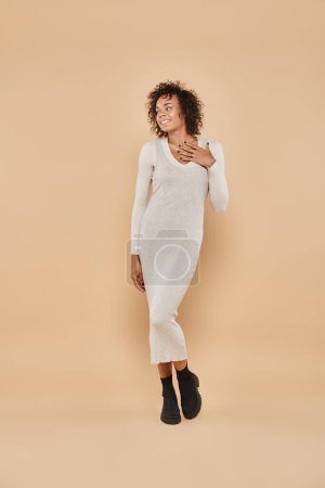 curly african american woman posing in midi dress and boots on beige backdrop, autumn fashion
