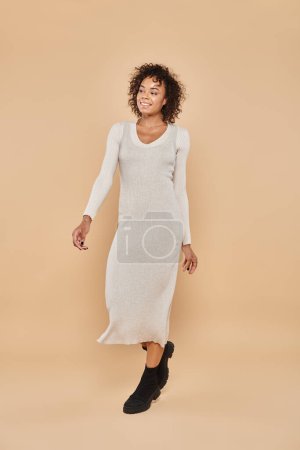 brunette african american woman standing in midi dress and boots on beige backdrop, autumn fashion