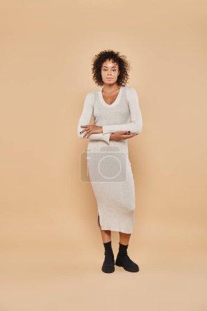 brunette african american woman standing in midi dress and boots on beige backdrop, autumn style