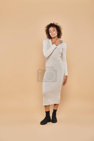 happy african american woman  in midi dress and boots adjusting curly hair on beige backdrop