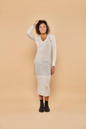 happy african american woman standing in knitted midi dress on beige backdrop, autumn style