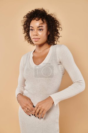 elegant african american woman standing in knitted white dress on beige backdrop, autumn fashion