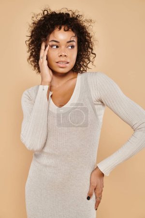 elegant african american woman in knitted dress and standing with hand on hip on beige backdrop