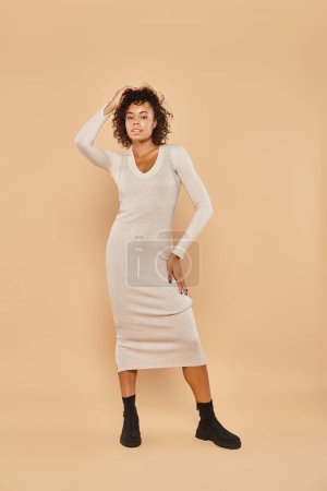 elegant african american woman standing in midi dress and boots on beige backdrop, autumn style