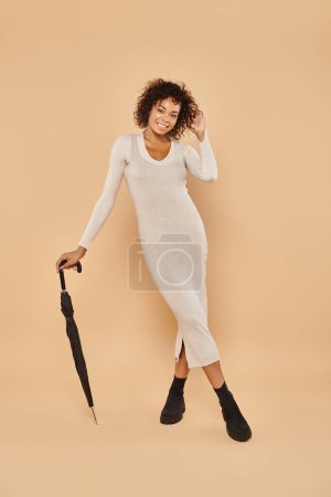 happy african american woman in midi dress and boots standing with umbrella on beige, autumn style