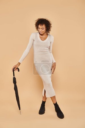 pleased african american woman in midi dress and boots standing with umbrella on beige, autumn style
