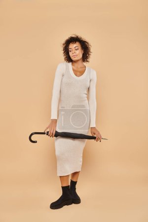 curly african american woman in midi dress and boots standing with umbrella on beige, autumn style