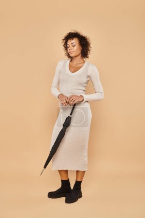 curly african american woman in midi dress and boots standing with umbrella on beige, autumn look