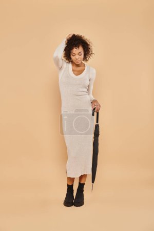 beautiful african american woman in midi dress and boots posing with umbrella on beige, autumn look