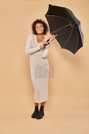 glad african american woman in midi dress and boots holding black umbrella on beige, autumn look