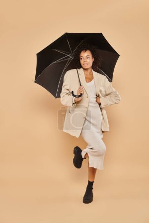 stylish african american woman in midi dress and autumnal blazer standing under umbrella on beige