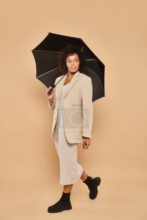 Photo for Curly african american woman in midi dress and autumnal blazer walking under umbrella on beige - Royalty Free Image