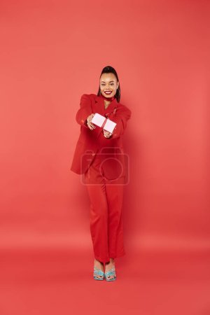 full length of african american woman posing in suit and holding Christmas present on red backdrop