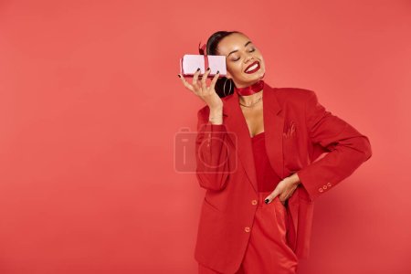 cheerful african american woman posing in red suit with hand on hip and holding Christmas present